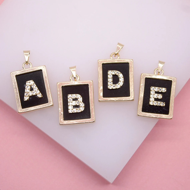 Four gold rectangular letter pendants featuring sparkling rhinestone initials A, B, D, and E against black backgrounds rest on a pink surface.