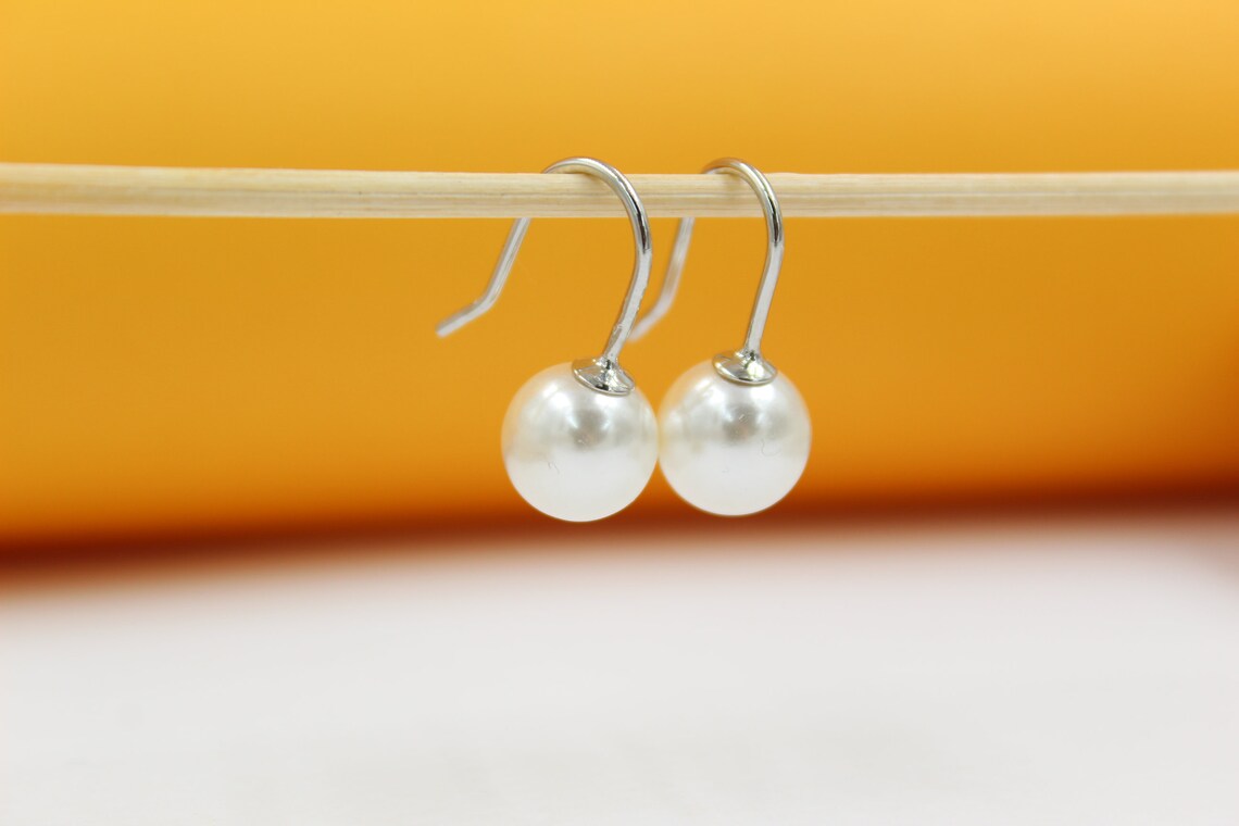 A pair of elegant silver pearl drop earrings hang neatly from a wooden stick against a vibrant orange backdrop.