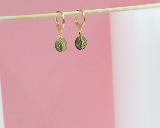 A pair of dainty gold huggie hoop earrings featuring dangling Saint Benedict medal charms hangs on a wooden stick against a pink and white background.