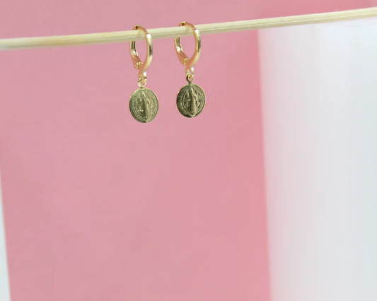 A pair of dainty gold huggie hoop earrings featuring dangling Saint Benedict medal charms hangs on a wooden stick against a pink and white background.