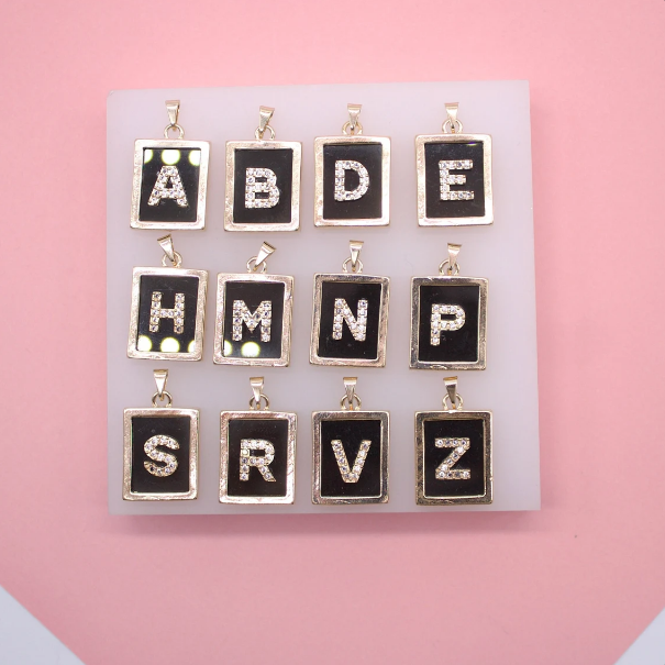 A white display board showcases twelve rectangular gold initial alphabet pendants featuring sparkly rhinestone letters set against a black background.