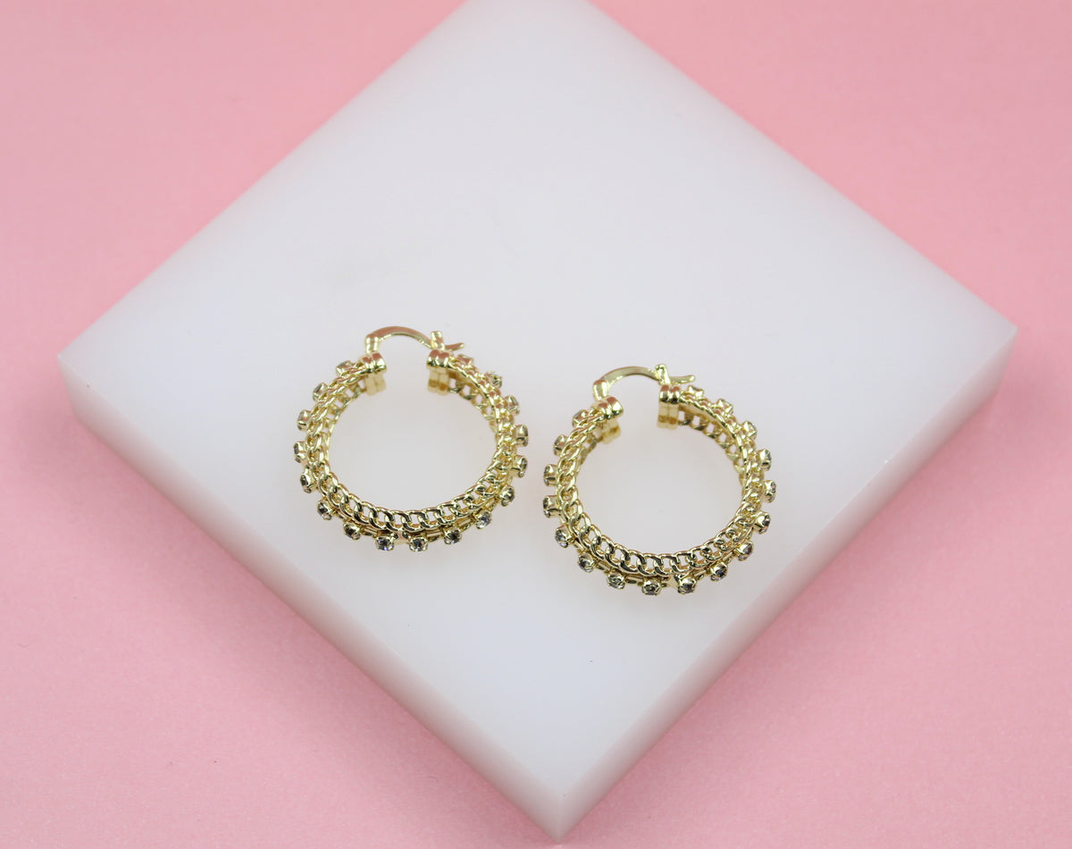 A pair of textured gold hoop earrings with small crystals rest on a white display block against a pink background.