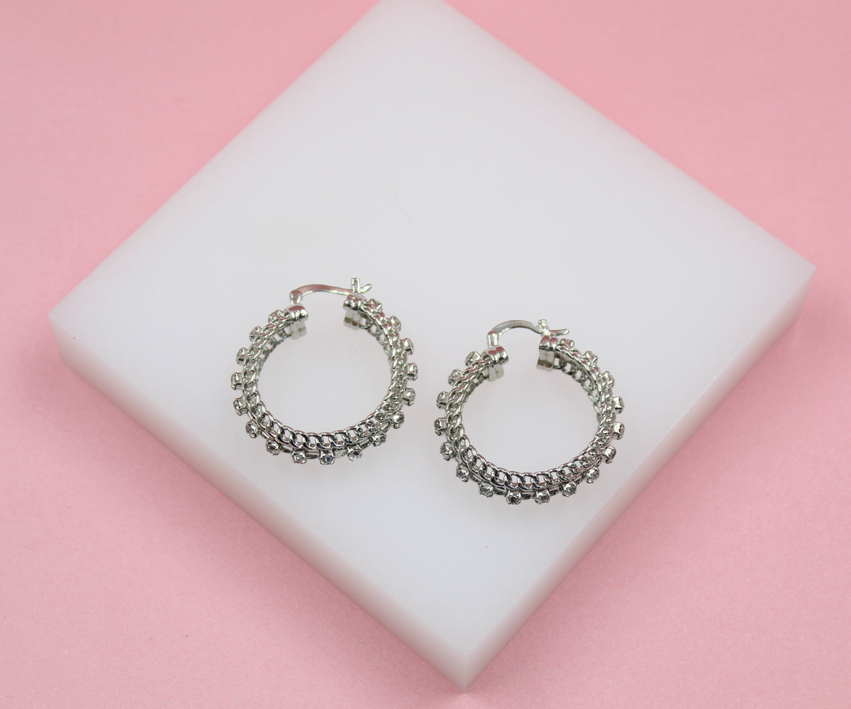 A pair of sparkly silver hoop earrings rests on a white display block against a soft pink background.