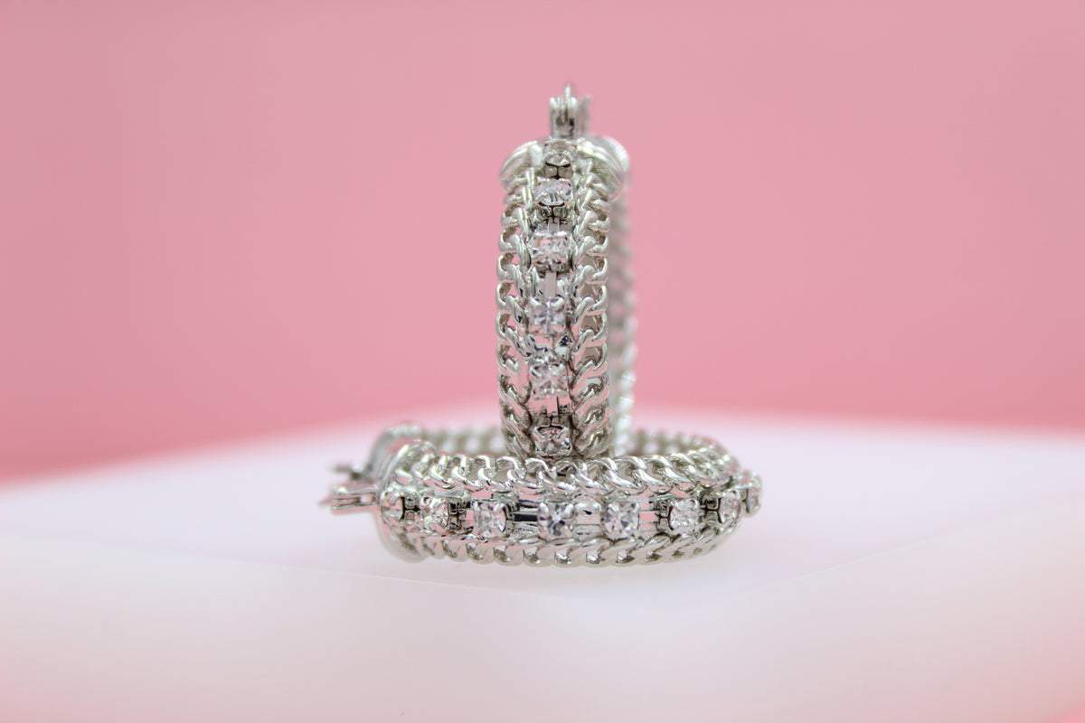 Two ornate silver hoop earrings encrusted with sparkling cubic zirconia stones rest against a soft pink backdrop.