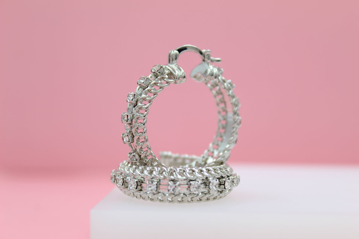 A pair of sparkling silver hoop earrings with intricate chain designs and embedded rhinestones rests on a white display stand against a soft pink background.