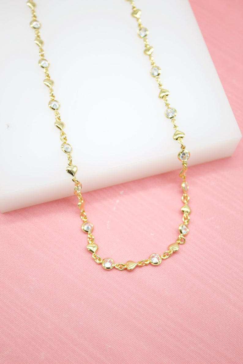 A delicate gold chain necklace with alternating heart charms and sparkling CZ crystals rests on a white display against a pink background.