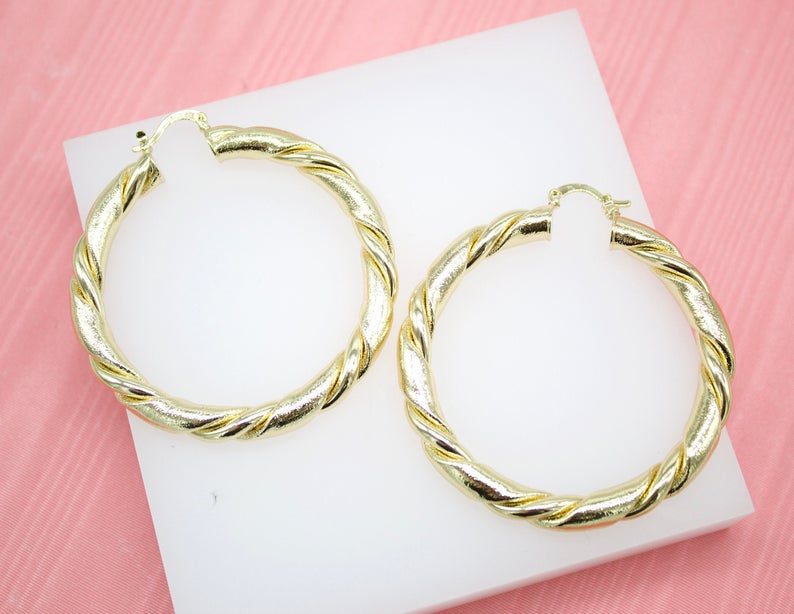 A pair of large, gold twist hoop earrings rests on a white display block against a pink background.