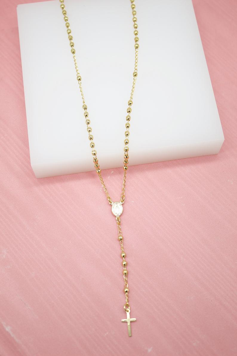 A gold beaded rosary necklace with a crucifix rests on a white display block over a pink surface.