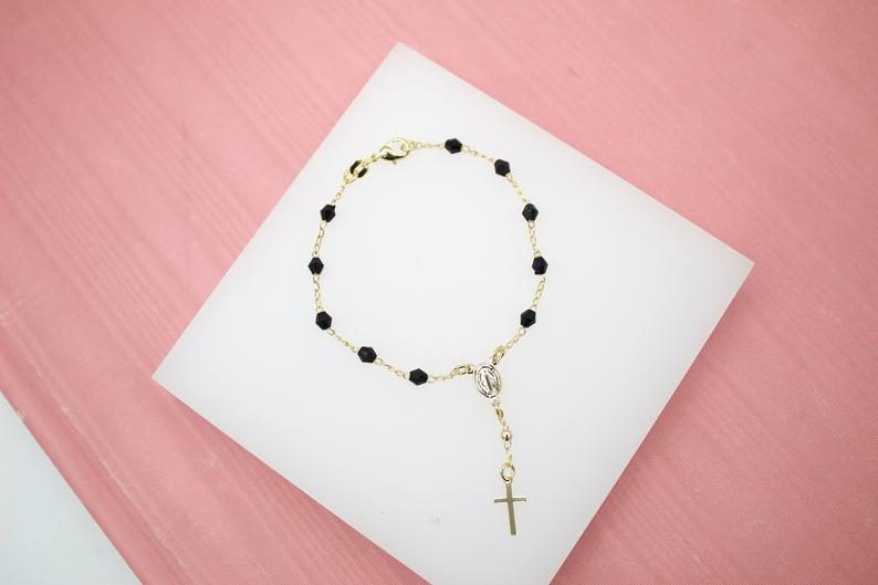 A gold rosary bracelet with black beads lies neatly on a white display square against a soft pink background.