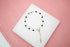 A gold rosary bracelet with black beads lies neatly on a white display square against a soft pink background.
