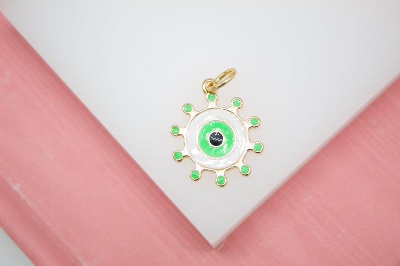 A gold-plated evil eye pendant features vibrant green enamel and white accents resting on a white display surface.