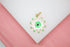 A gold-plated evil eye pendant features vibrant green enamel and white accents resting on a white display surface.