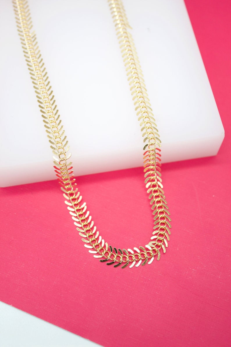 A gold leaf-pattern chain necklace rests on a white display block against a bright pink background.