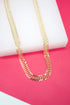 A gold leaf-pattern chain necklace rests on a white display block against a bright pink background.