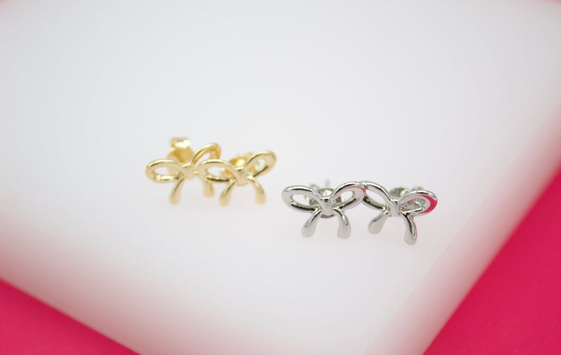 A pair of gold bow stud earrings rests alongside a matching pair of silver bow stud earrings on a white and pink display surface.
