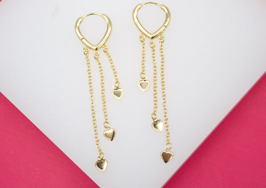 A pair of gold heart-shaped hoop earrings featuring dangling chains with tiny heart charms rests on a white display surface.