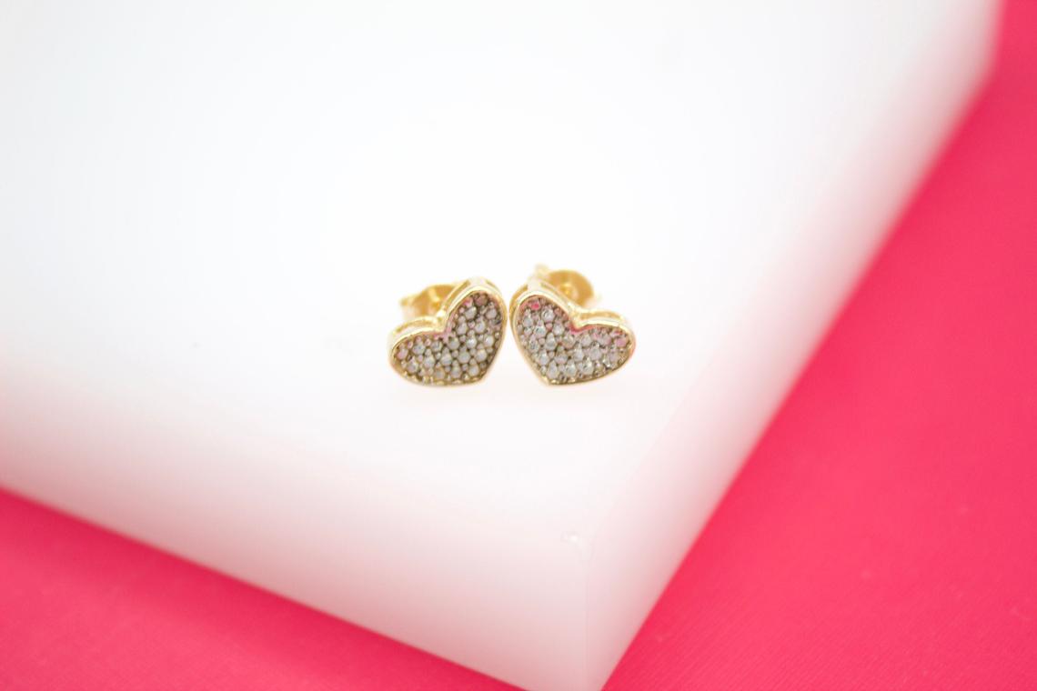 A pair of gold heart-shaped stud earrings sparkle with tiny embedded crystals on a white display block.