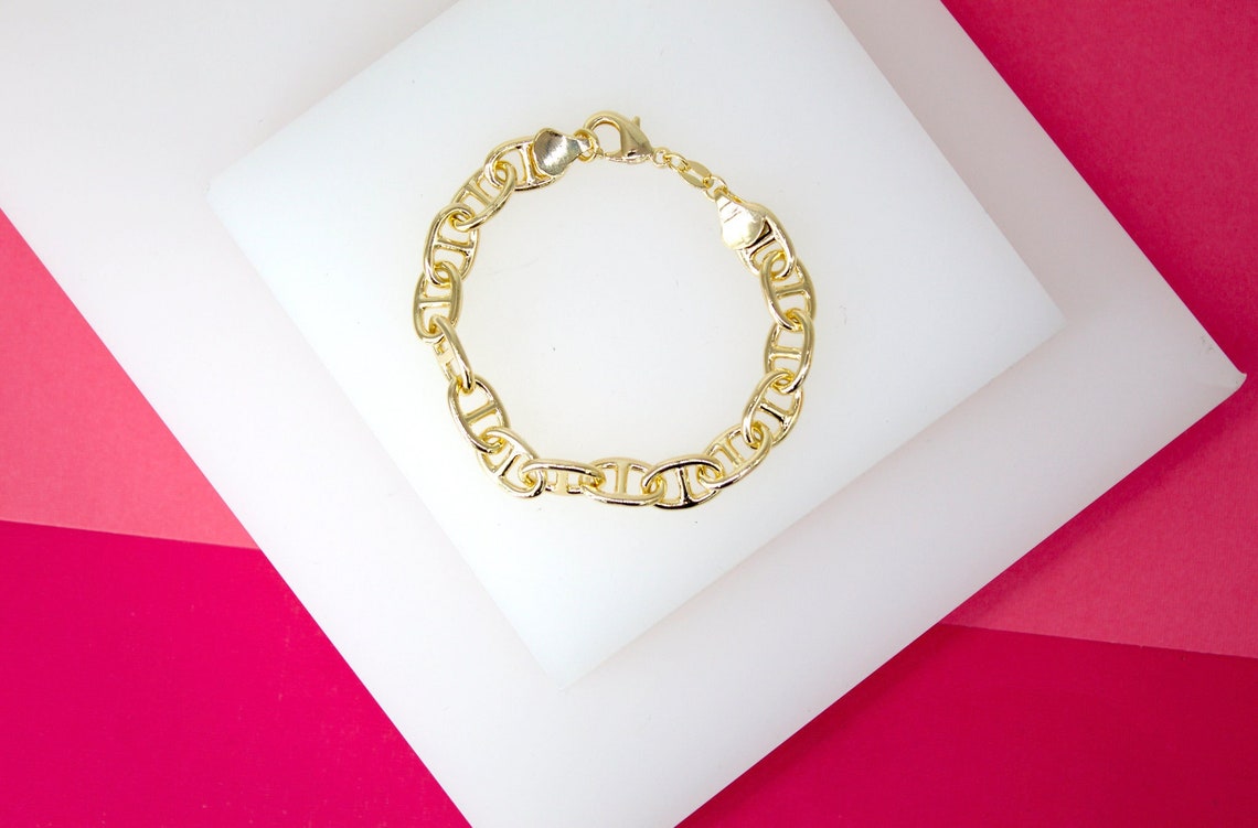A glossy gold mariner link bracelet rests neatly on a white display block against a vibrant pink background.