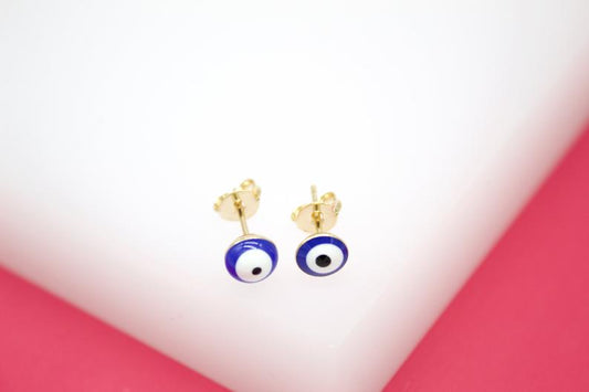 A pair of gold evil eye stud earrings rests on a white and pink display stand.