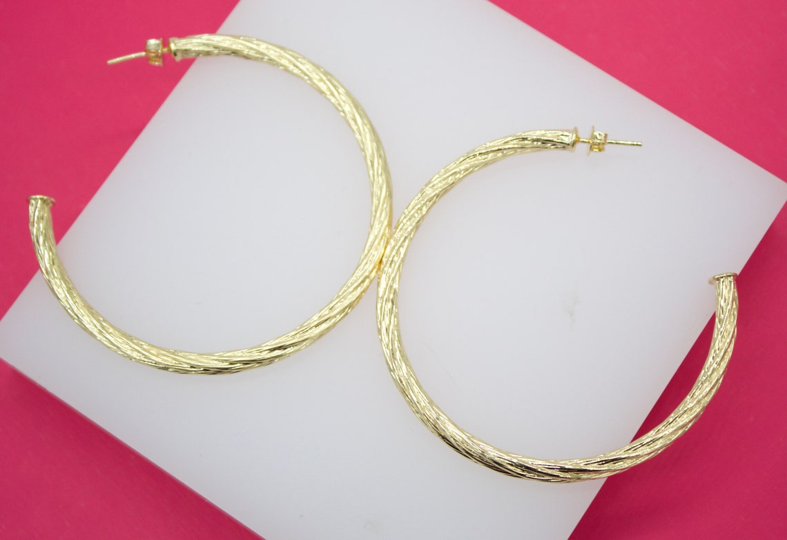 A pair of large, textured gold hoop earrings rests on a white display surface against a vibrant pink background.