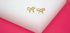 A pair of polished gold bow-shaped stud earrings rest neatly on a white display block against a vibrant pink background.
