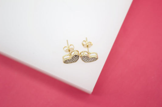 A pair of gold heart-shaped stud earrings encrusted with small pave diamonds rests on a white display surface against a pink background.