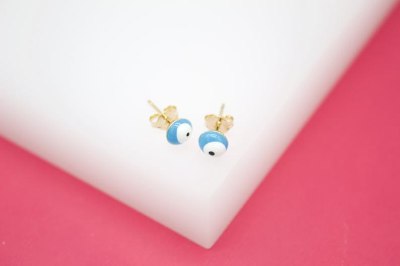 A pair of gold evil eye stud earrings with blue enamel rests on a white and pink display.