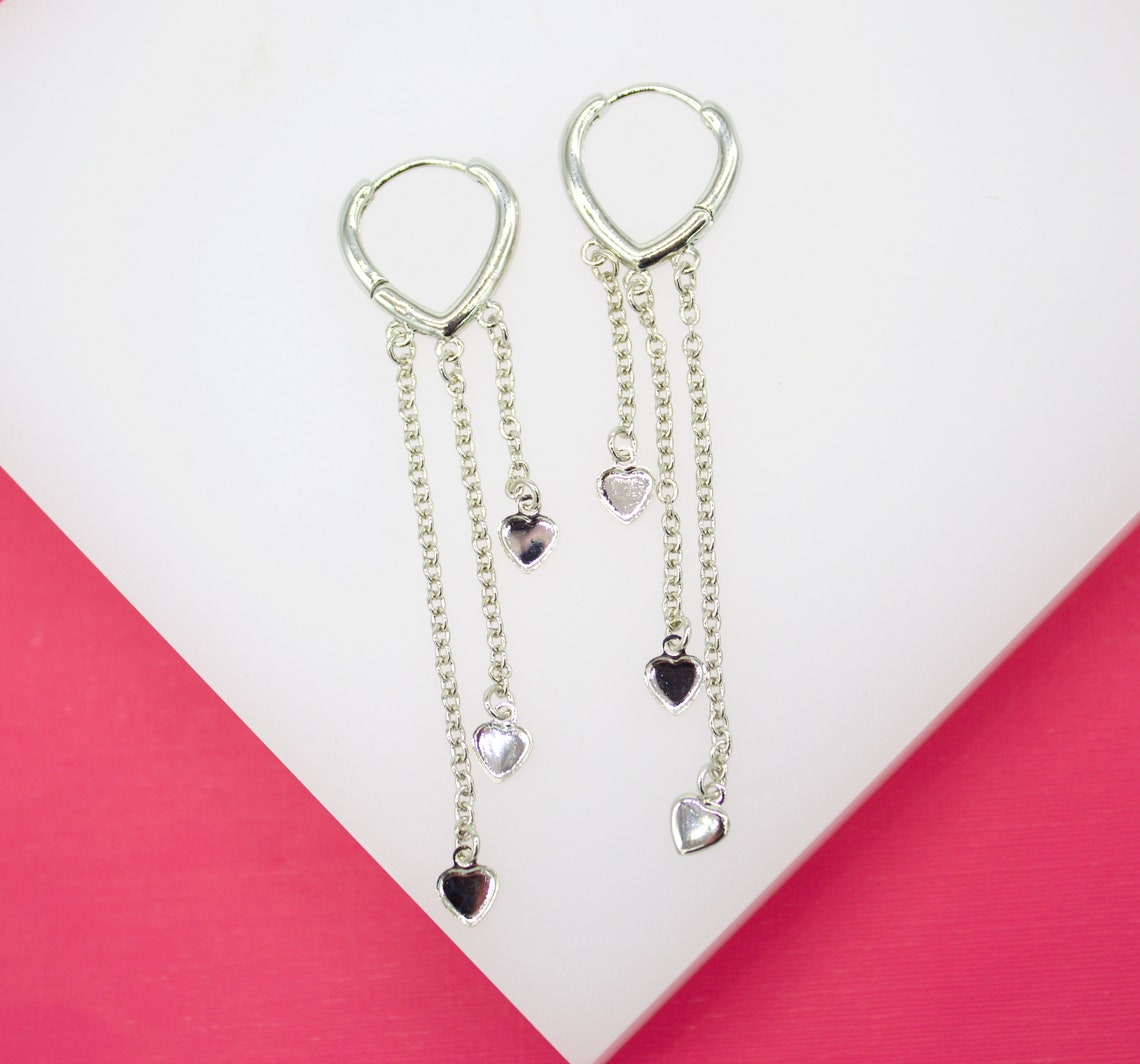 A pair of dangling silver heart hoop earrings rests against a white and pink display surface.