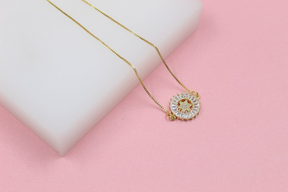 A delicate gold necklace features a sparkling sunburst pendant with baguette-cut crystals and a central star motif, resting against a white and pink background.