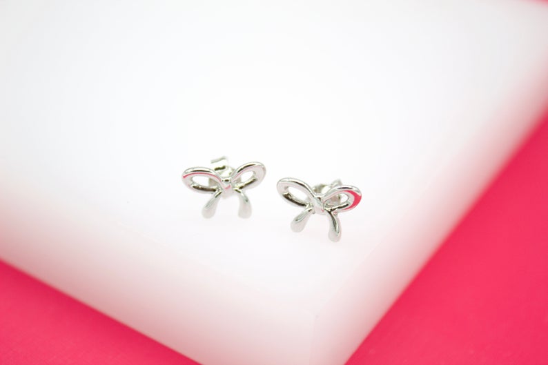 A pair of dainty silver bow stud earrings rest on a white display stand against a vibrant pink background.