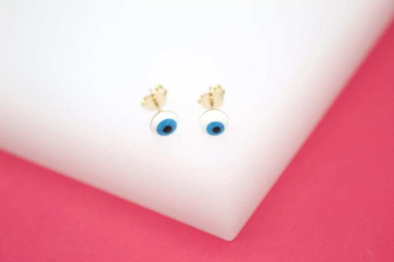 A pair of gold evil eye stud earrings rests on a white display against a pink background.