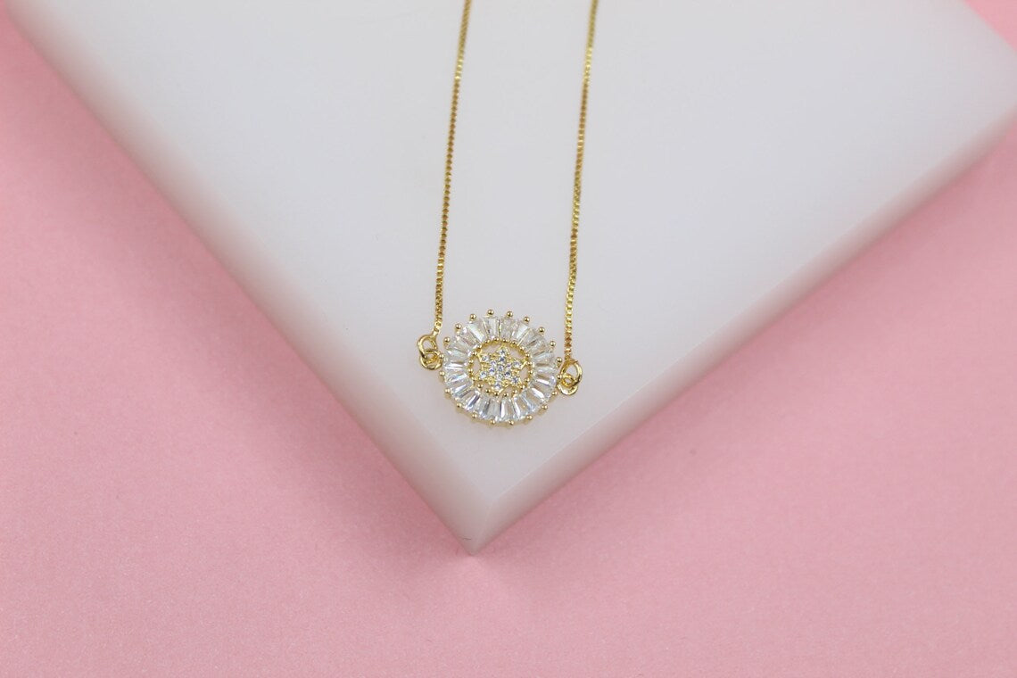 A delicate gold chain necklace with a circular pendant featuring sparkling baguette and round cubic zirconia stones rests on a white display against a pink background.