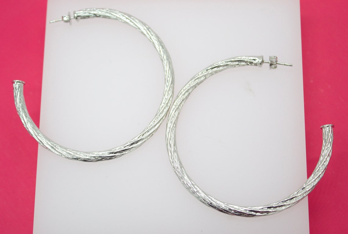 A pair of large silver textured hoop earrings rests on a white display surface against a bright pink background.