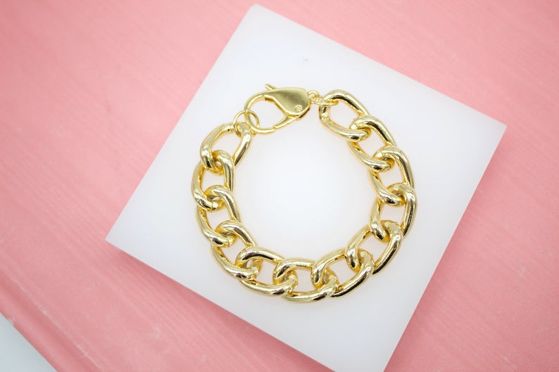 A chunky gold link bracelet rests on a white square display against a pink background.