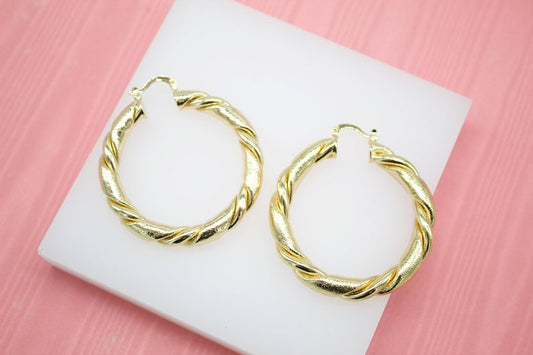 A pair of thick gold twist hoop earrings rests on a white display stand against a pink background.