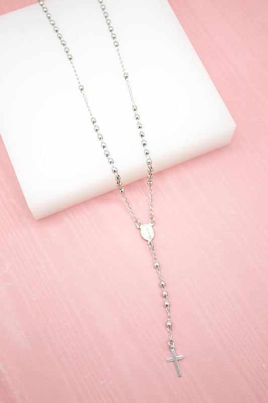 A gleaming silver rosary necklace rests elegantly on a white display block against a soft pink background.