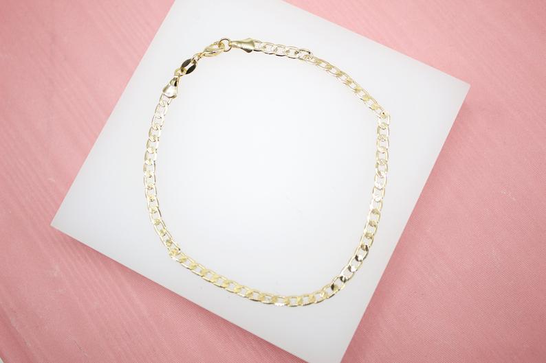 A delicate gold chain anklet rests on a white display card against a pink background.