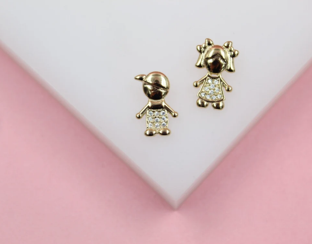 A pair of gold earrings shaped like a boy wearing a cap and a girl with pigtails, both featuring crystal-embellished shirts, rests on a white display against a pink background.