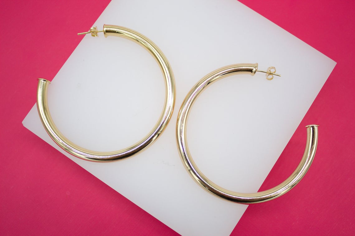 A pair of large, polished gold-plated hoop earrings rests on a white display block against a vibrant pink background.