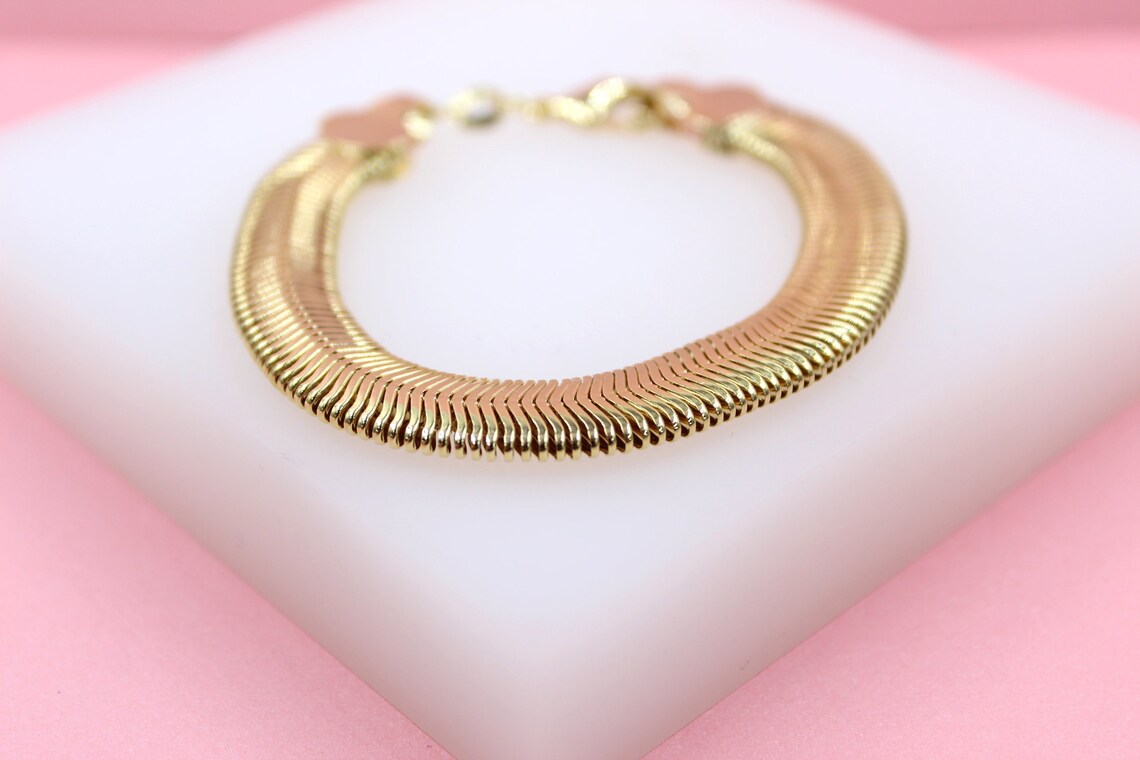 A gold herringbone chain bracelet rests on a white display stand against a soft pink background.