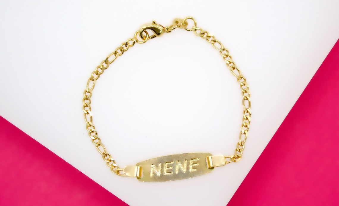 A gold ID bracelet rests on a white and pink background, displaying the engraved name "NENE" on its central plaque.