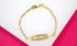 A gold ID bracelet rests on a white and pink background, displaying the engraved name "NENE" on its central plaque.