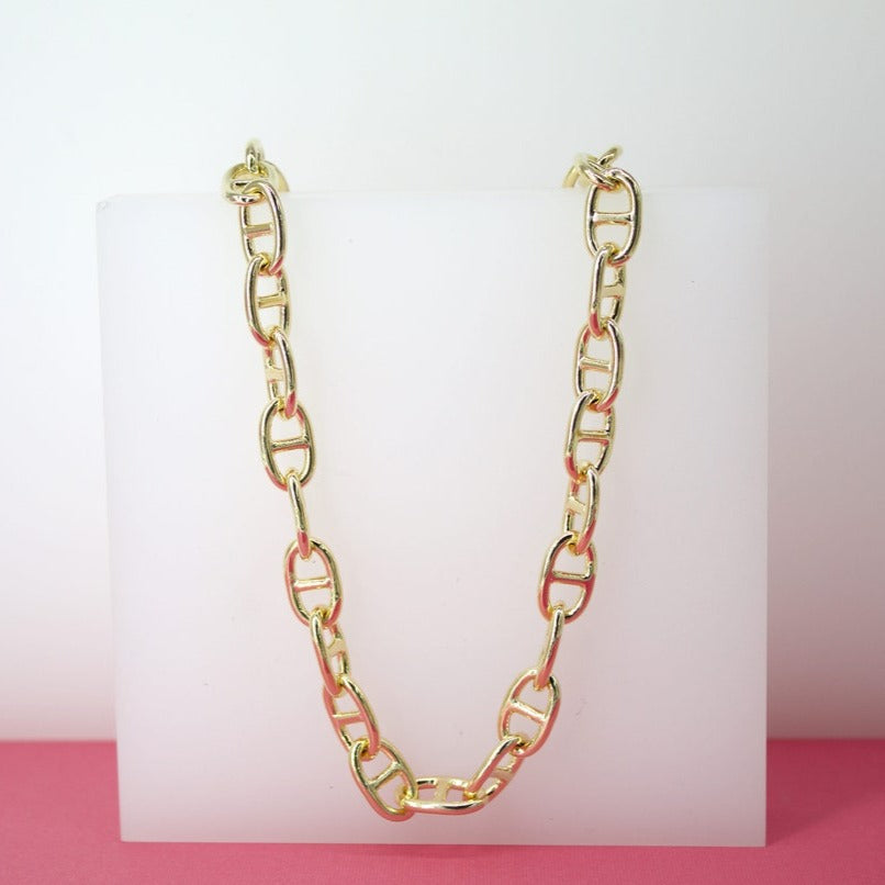 A chunky gold chain-link necklace rests gracefully on a white display block against a pink surface.