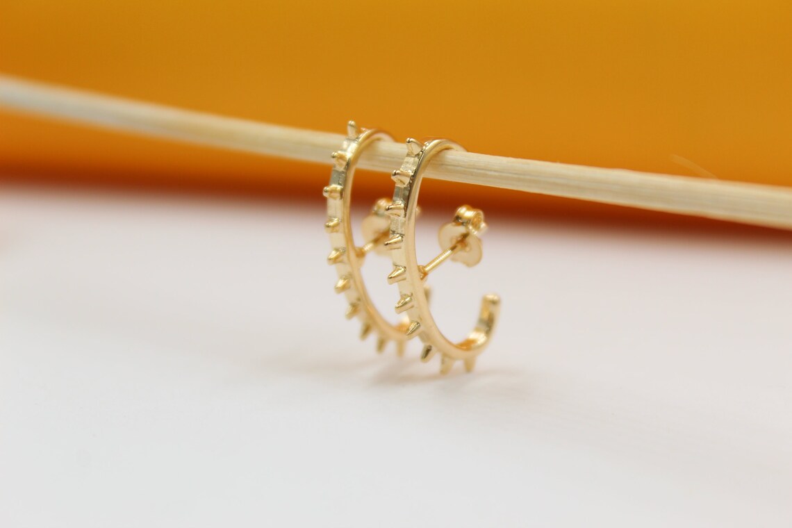 A pair of gold spike hoop earrings hangs delicately from a wooden stick against a soft-focus background.
