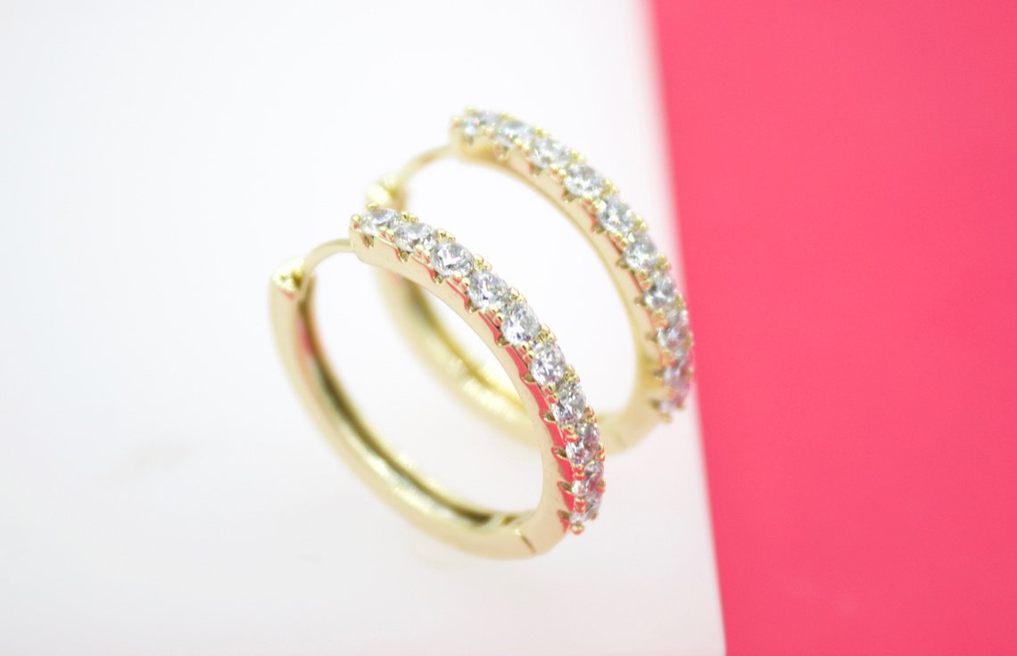 A pair of sparkling gold hoop earrings encrusted with clear cubic zirconia stones rests on a white surface against a bright pink background.