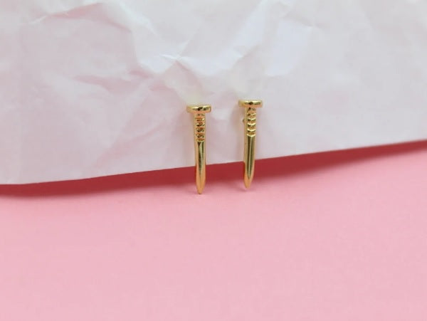 Two dainty gold nail-shaped stud earrings rest neatly against a pink and white textured backdrop.