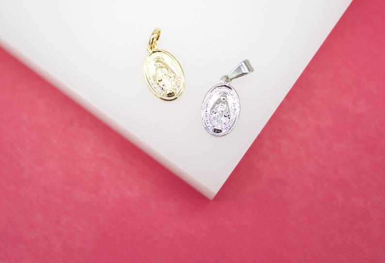 A gold-plated and a silver-plated Virgin Mary miraculous medal pendant rest side-by-side on a white display against a vibrant pink background.