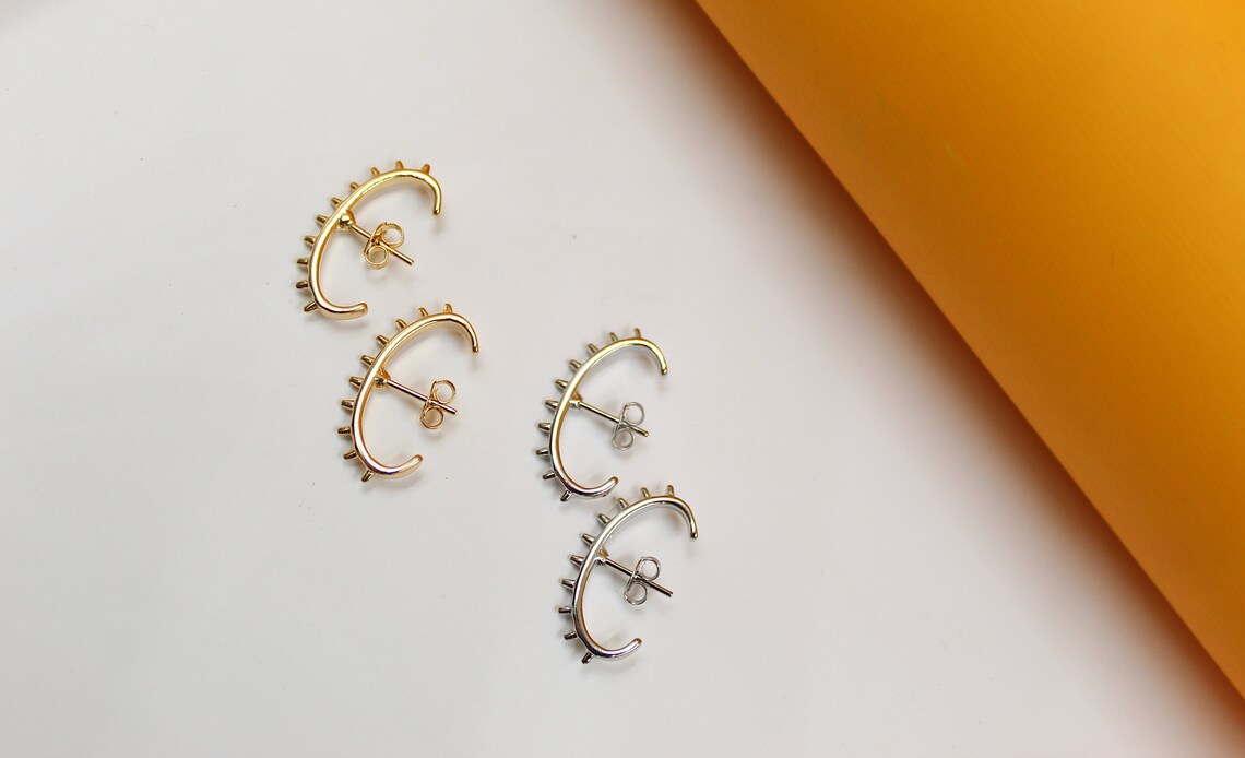 Two pairs of gold and silver spiky ear cuff earrings rest on a white surface next to an orange roll.