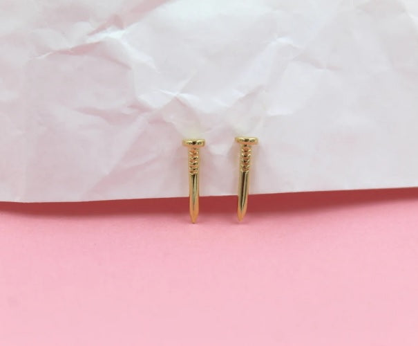 Two gold nail-shaped stud earrings rest against a backdrop of white crinkled paper and a soft pink surface.