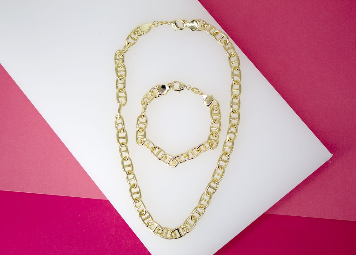 A matching gold mariner chain necklace and bracelet set rests on a white display against a pink background.